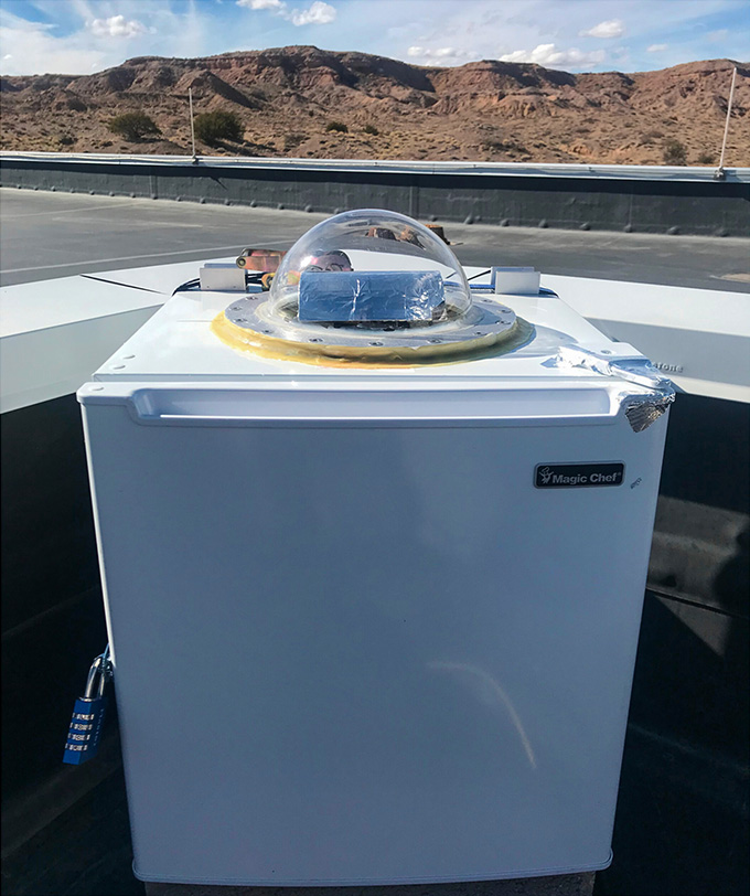 A mini-fridge with a clear dome on top sits against a desert landscape